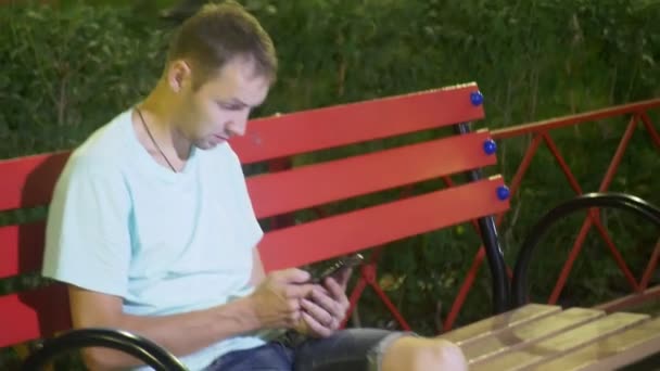 Привлекательный грустный мужчина сидит на скамейке со смартфоном. поздно теплым летним вечером. в парке. 4k, steady icam shot — стоковое видео