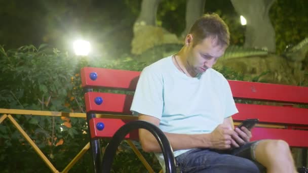 有魅力的可悲男人坐在一台智能手机的长凳上。在一个温暖的夏日夜晚的深夜。在公园里。4k, 替身射击 — 图库视频影像