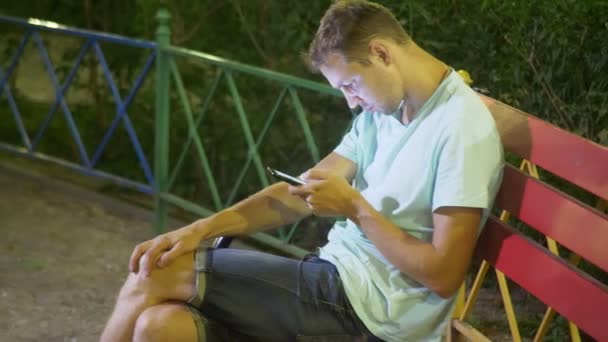 Homem triste atraente sentado em um banco com um smartphone. tarde em uma noite quente de verão. no parque. 4K, steadicam tiro — Vídeo de Stock