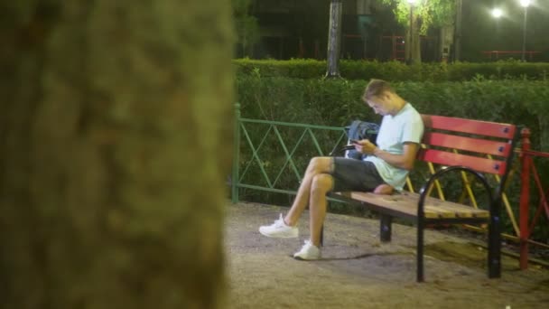 Aantrekkelijke trieste man zittend op een bankje met een smartphone. laat op een warme zomeravond. in het park. 4k, steadicam schot — Stockvideo