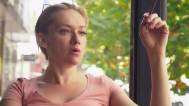 Genç bir kadın bir güneşli yaz sabahı bir kafede bir verandada otururken ince sigara içiyor. 4k, ağır çekim