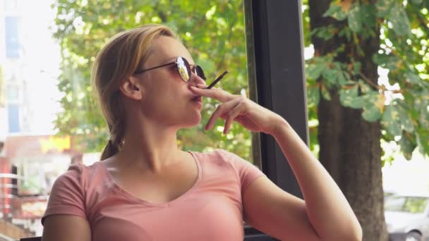 Una joven fuma cigarrillos finos mientras está sentada en una terraza en un café, en una soleada mañana de verano. 4k, cámara lenta — Vídeo de stock