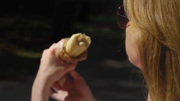 Девушка в солнечных очках, с удовольствием ест хот-доги, сидит в городском парке на скамейке. 4k, slow motion — стоковое видео