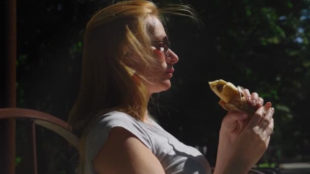 Chica en gafas de sol, comer perritos calientes con placer, sentado en un parque de la ciudad en un banco. 4k, cámara lenta — Vídeos de Stock