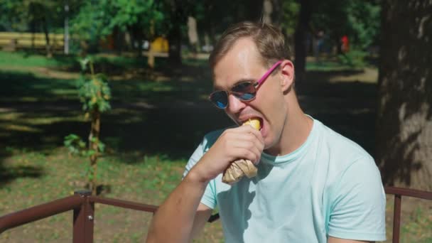 Tipo con gafas de sol, comiendo perritos calientes con placer, sentado en un parque de la ciudad en un banco. 4k, cámara lenta — Vídeo de stock