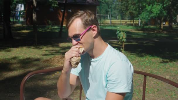 Facet w okulary, jedzenie hot dogi z przyjemnością, siedząc na ławce w parku. 4k, zwolnionym tempie — Wideo stockowe