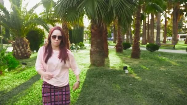 Jovem bela menina ruiva dançando no parque. mulher em uma mini saia dançando em um parque tropical em um fundo de palmas. 4k, câmera lenta — Vídeo de Stock