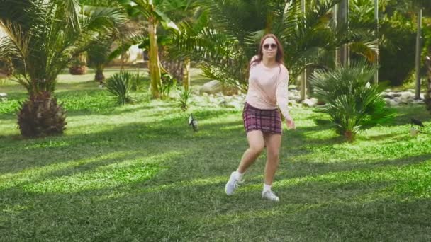 Όμορφη κοκκινομάλλα κοπέλα χορεύει στο πάρκο. γυναίκα σε μια μίνι φούστα χορού σε ένα τροπικό πάρκο σε ένα φόντο από φοίνικες. 4k, αργή κίνηση — Αρχείο Βίντεο