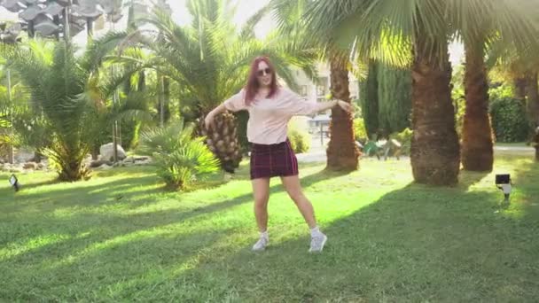 Jovem bela menina ruiva dançando no parque. mulher em uma mini saia dançando em um parque tropical em um fundo de palmas. 4k, câmera lenta — Vídeo de Stock