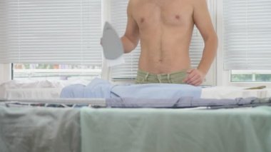 ev işi sırasında Gömlek Ütü kurulu dumanı tüten demir, kullanarak arkasında duran çıplak gövde olan genç adam. 4k, ağır çekim