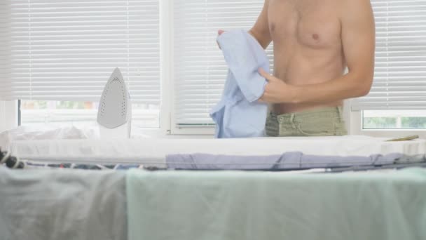 Молодий чоловік з голим торсом стоїть за дошкою, використовуючи парову праску, прасувальну сорочку під час домашніх завдань. 4k, повільний рух — стокове відео