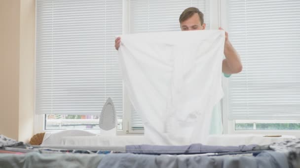 Joven, parado detrás de la tabla usando plancha humeante, camisa de planchar durante las tareas domésticas. 4k, cámara lenta — Vídeo de stock