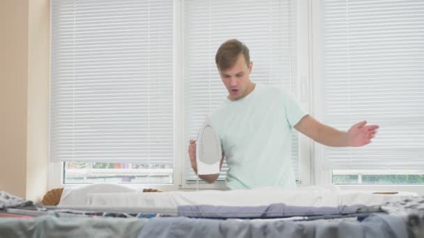 Молодий чоловік, стоїть за дошкою з використанням парового заліза, прасувальної сорочки під час домашніх завдань. 4k, повільний рух — стокове відео