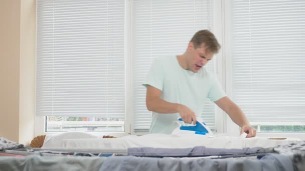 Junger Mann, der mit dampfenden Bügeleisen hinter Bord steht und während der Hausarbeit Hemd bügelt. 4k, Zeitlupe — Stockvideo