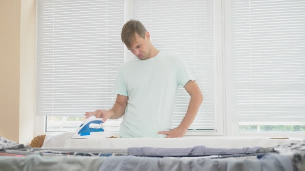Молодой человек, стоящий за бортом, используя дымящийся утюг, гладильная рубашка во время работы по дому. 4k, slow-motion — стоковое видео