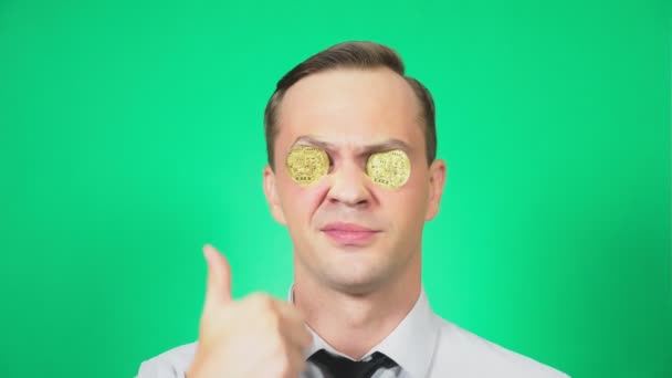 Portrait d'un beau jeune homme avec une pièce Bitcoin devant les yeux. Il montre un pouce levé, un super signe. Concentre-toi sur le visage. fond vert, 4k, ralenti — Video