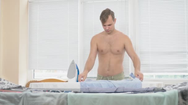 家事の中にシャツをアイロンのスチーム アイロンを使用してボードの後ろに裸の胴体立って若い男。4 k、スローモーション — ストック動画
