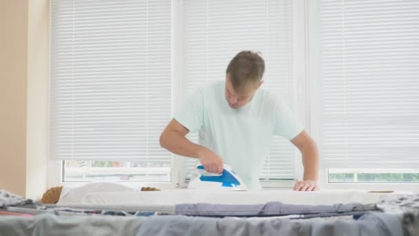 Молодой человек, стоящий за бортом, используя дымящийся утюг, гладильная рубашка во время работы по дому. 4k, slow-motion — стоковое видео