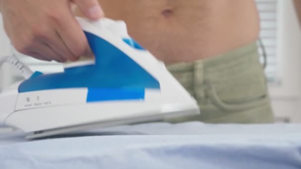 Joven con torso desnudo parado detrás de la tabla usando plancha humeante, camisa de planchar durante las tareas domésticas. 4k, cámara lenta — Vídeo de stock