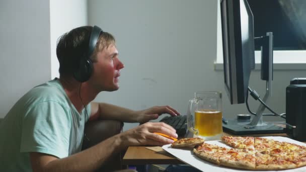 Технологии, игры, развлечения, игра и концепция людей. Молодой человек играет в компьютерную игру дома, пьет пиво и ест пиццу. 4k, slow motion — стоковое видео