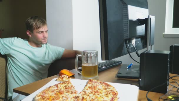 A satiated man sits on the couch at home, drinks beer and eats pizza, using the Internet on a computer, watching an online movie or working as an independent freelancer. 4k, slow motion — Stock Video