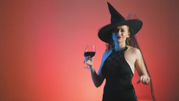 Mooie jonge sexy vrouw in een zwarte heks kostuum en hoed, met een glas wijn kijkt in de camera en glimlach. Kopieer ruimte. 4k, slow-motion — Stockvideo