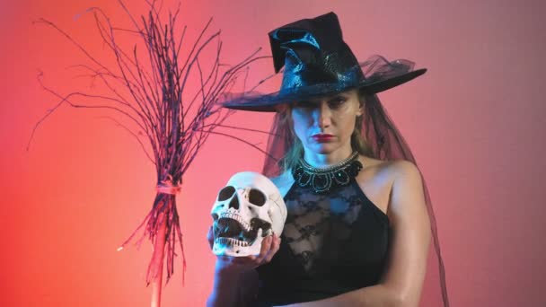 Beautiful young sexy woman in a black witch costume and hat, with a broom and a skull in her hands. copy space. 4k, slow motion — Stock Video