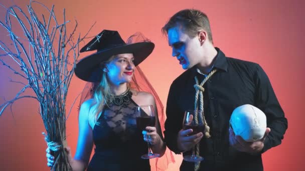 Um belo casal, um homem e uma mulher em trajes de bruxa e zumbi Halloween, 4K, tiro em movimento lento, segurando copos de vinho e um crânio. parabéns pelo feriado — Vídeo de Stock
