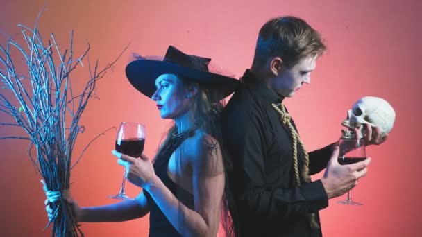 Um belo casal, um homem e uma mulher em trajes de bruxa e zumbi Halloween, 4K, tiro em movimento lento, segurando copos de vinho e um crânio. parabéns pelo feriado — Vídeo de Stock