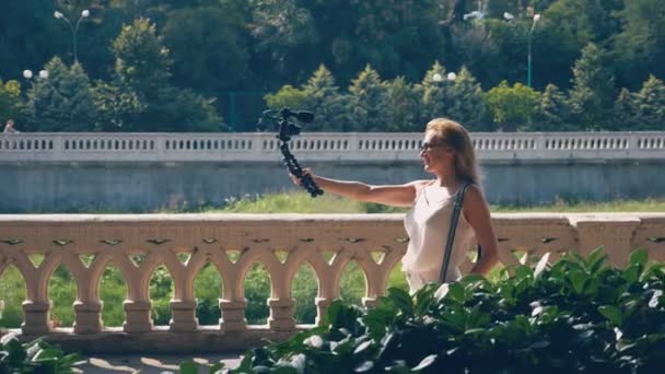 Vackra lycklig kvinna bloggare med en kamera, leder hennes reportage strosa längs banvallen av resort staden. vinden utvecklar hennes hår, solljus glintes genom grenarna i träden — Stockvideo