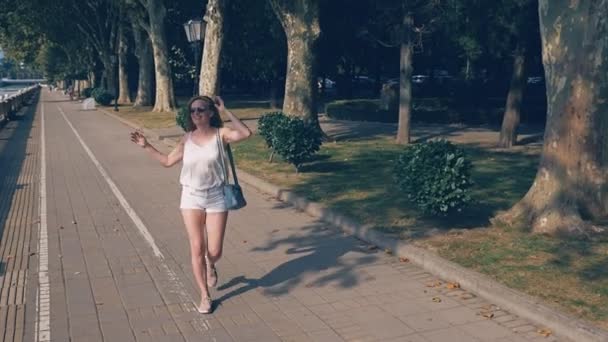 Красивая счастливая женщина блоггер с камерой, ведет свой репортаж прогуливаясь по набережной курортного города. Ветер развивает ее волосы, солнечный свет сверкает сквозь ветви деревьев — стоковое видео
