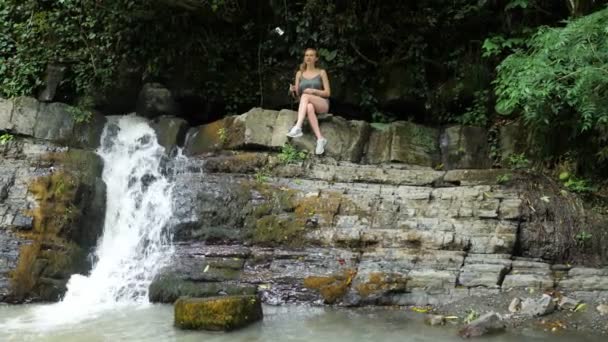 Blogueira está liderando um relatório sentado em uma rocha perto da cachoeira. A natureza pitoresca de uma bela cachoeira e uma esmeralda de um lago de água doce no ambiente florestal de selvas selvagens. 4k — Vídeo de Stock