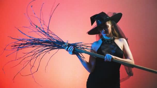 Красивая молодая сексуальная женщина в чёрном костюме ведьмы и шляпе, с метлой, смотрящей в камеру и улыбающейся. скопировать пространство. 4k, slow motion — стоковое видео