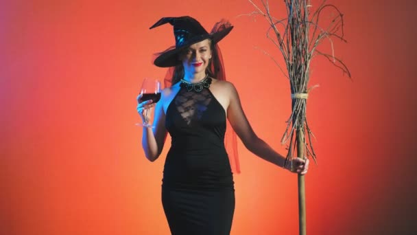 Όμορφη νεαρή γυναίκα σέξι σε μια μάγισσα μαύρο κοστούμι και καπέλο, με ένα ποτήρι κρασί και μια σκούπα κοιτάζει επίμονα στη φωτογραφική μηχανή και χαμόγελα. χώρο αντίγραφο. 4k, αργή κίνηση — Αρχείο Βίντεο