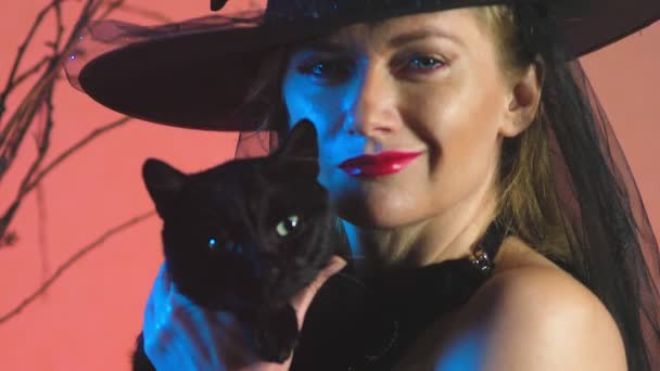 Belle jeune femme sexy dans un costume de sorcière noire et chapeau, avec un balai et un chat noir dans ses mains regardant dans la caméra et souriant. espace de copie. 4k, au ralenti — Video