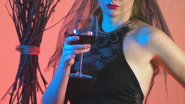 Beautiful young sexy woman in a black witch costume and hat, with a glass of wine and a broom stares into the camera and smiles. copy space. 4k, slow motion — Stock Video