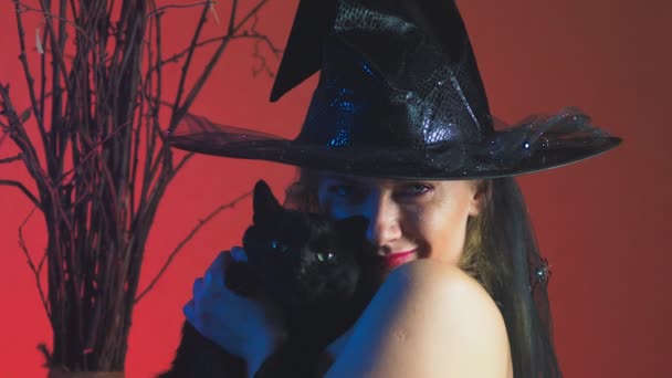 Mooie jonge sexy vrouw in een zwarte heks kostuum en hoed, met een bezem en een zwarte kat in haar handen op zoek naar de camera en glimlachen. Kopieer ruimte. 4k, slow-motion — Stockvideo