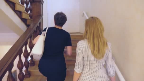 Zwei schlanke Frauen, die Mutter und ihre erwachsene Tochter, steigen die Holztreppe mit Geländern hinauf, drinnen. 4k, — Stockvideo