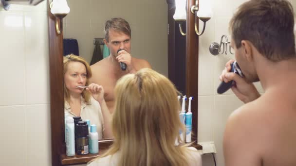 Hermosa pareja, hombre y mujer, lavar juntos en el baño delante del espejo. 4k, cámara lenta, una mujer se cepilla los dientes, un hombre se afeita . — Vídeos de Stock