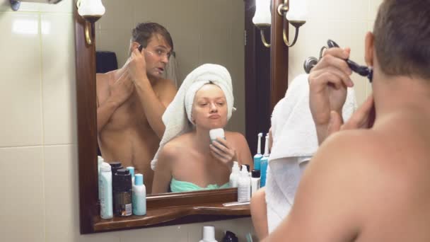 Vackert par, man och kvinna, tvätta tillsammans i badrummet framför spegeln. 4k, ultrarapid, de man rakar ansiktet med en verktygsmaskin, en kvinna gör en epilering av håret på henne — Stockvideo