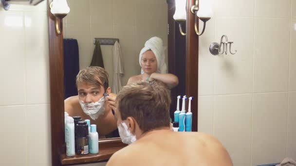 Hermosa pareja, hombre y mujer, lavar juntos en el baño delante del espejo. 4k, cámara lenta, una mujer se cepilla los dientes, un hombre se afeita . — Vídeos de Stock