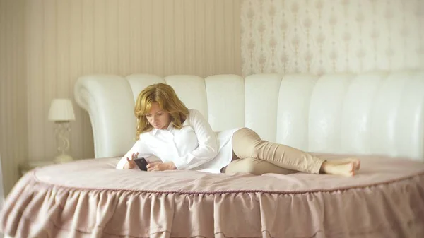 Een trendy dame gekleed in een witte blouse en een beige broek. Een jonge vrouw maakt gebruik van een mobiele telefoon binnenshuis met een prachtige interieur. — Stockfoto