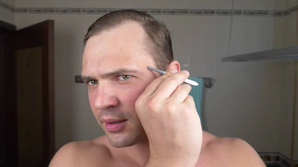 A handsome man pulls his hair out of his eyebrows with tweezers in front of a mirror in the bathroom. Eyebrow epilation. — Stock Photo, Image