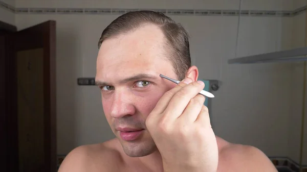 Een knappe man trekt zijn haar uit zijn wenkbrauwen met pincet voor een spiegel in de badkamer. Epileren van de wenkbrauw. — Stockfoto