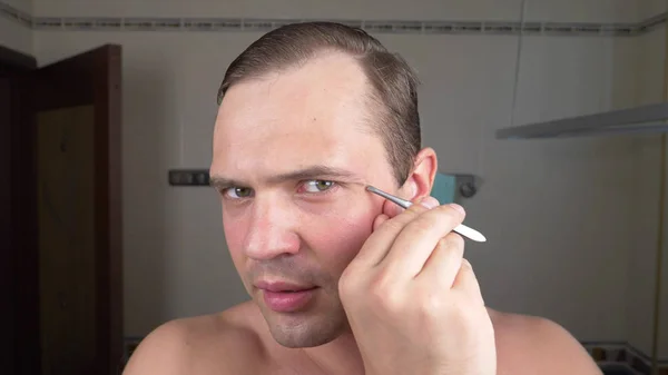 En stilig man drar sitt hår ur ögonbrynen med en pincett framför en spegel i badrummet. Ögonbryn epilering. — Stockfoto