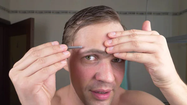 En stilig man drar sitt hår ur ögonbrynen med en pincett framför en spegel i badrummet. Ögonbryn epilering. — Stockfoto