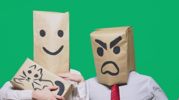 El concepto de emociones y gestos. Dos personas en bolsas de papel con emoticonos. Sonriente agresivo jura. La segunda le sonríe y acaricia al gato pintado en la caja . — Vídeos de Stock