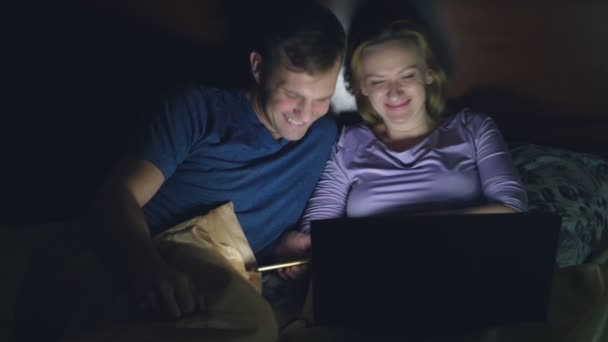 Coppia, uomo e donna, guardando un film su un computer portatile su un letto in camera da letto prima di andare a letto. guardando un film comico, il pubblico ride . — Video Stock