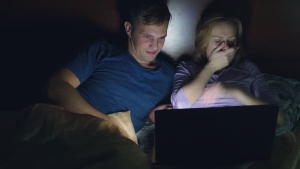 Par, man och kvinna, titta på en film på en bärbar dator på en säng i sovrummet innan sängen. Titta på en skräckfilm, få tittare rädd och äcklad — Stockvideo