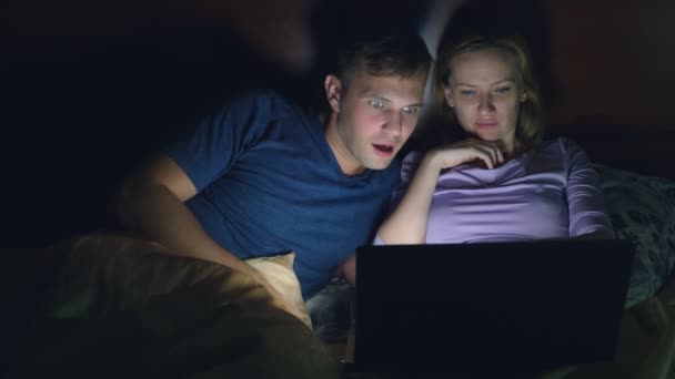 Paar, man en vrouw, lettend op een film op een laptop op een bed in de slaapkamer voor het slapen. kijken naar een spannende film, is het publiek kijken met belangstelling, verschillende emoties ervaren. — Stockvideo